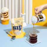 Loose-leaf Cinnamon Roll herbal latte blend by Sip Herbals shown with a French press pouring into a glass beside cinnamon rolls and a candle for a cozy aesthetic